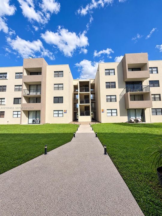Multi-story exterior featuring a concrete-finished facade, multiple private balconies with metal railings, and a paved walkway lined with bollard lighting