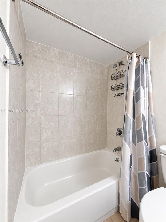 Full-size bathtub featuring beige wall tile surround, stainless steel shower fixtures, and a wall-mounted metal storage rack