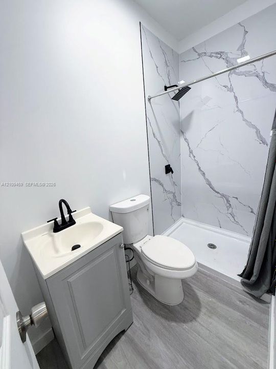 Bathroom featuring wood-finish flooring, a vanity with a drop-in sink, matte black faucet hardware, and a walk-in shower with stone-patterned wall panels and a rainfall showerhead