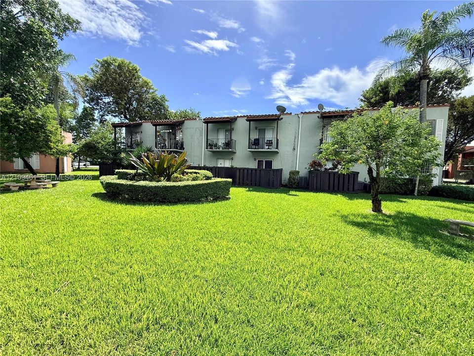 Expansive green space featuring mature trees, manicured hedges, building exteriors with terracotta-style roof accents, and private balconies
