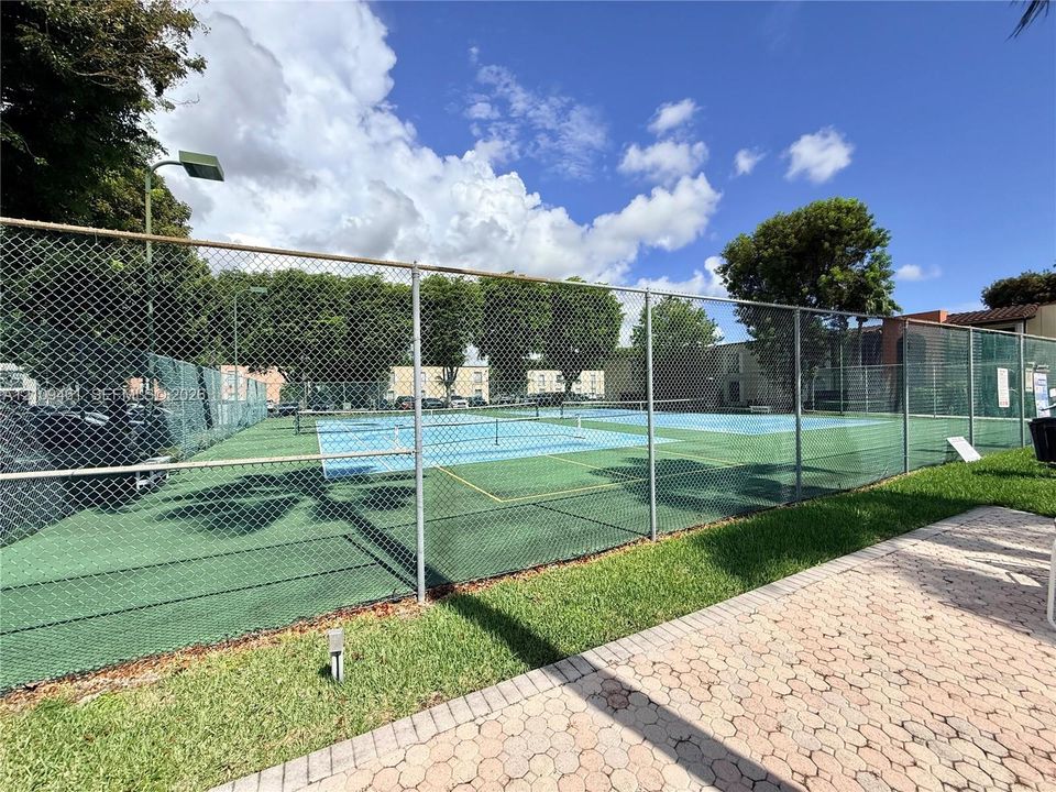 Hard-surfaced tennis courts featuring chain-link fencing, dual-tone court surfacing, and interlocking paver walkways