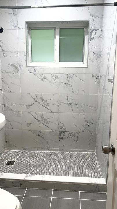 Walk-in shower enclosure featuring large-format veined tile surround, hex-pattern mosaic floor tiling, integrated horizontal window, and dark-finish square shower drain