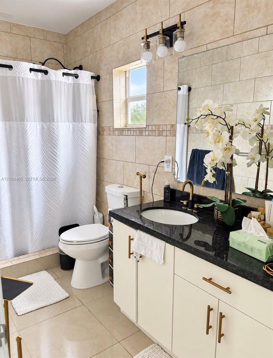 Bathroom featuring a vanity with dark stone countertop, undermount sink, brass-finish hardware, three-light fixture, and ceramic tile wall finishes