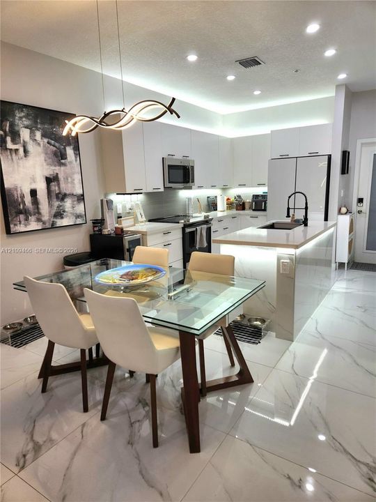 Kitchen featuring white flat-panel cabinetry, light-toned stone countertops, a central island with integrated under-counter lighting, large-format veined flooring, and modern sculptural pendant lighting