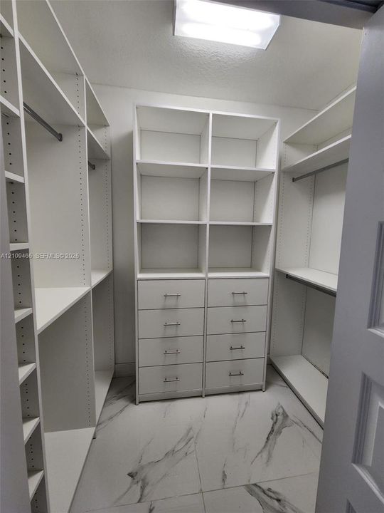 Walk-in closet featuring white built-in shelving, integrated dresser drawers with metallic hardware, hanging rods, and large-format porcelain tile flooring