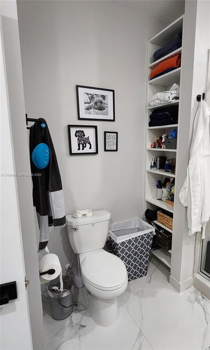 White ceramic toilet, built-in floor-to-ceiling white shelving, stone-finish flooring, and wall-mounted matte black hardware