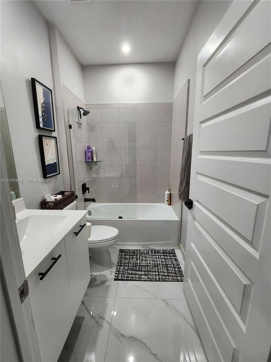 Bathroom featuring white cabinetry with matte black hardware, large-format tile flooring with marble-finish veining, a deep-set soaking tub, and a glass partition wall