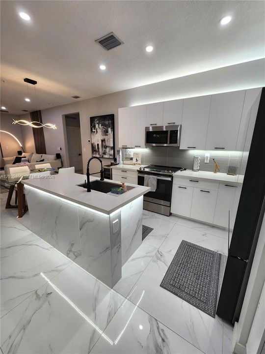 Kitchen featuring white flat-panel cabinetry, a central island with a stone-finish countertop, stainless steel appliances, integrated under-cabinet lighting, and large-format rectified tile flooring
