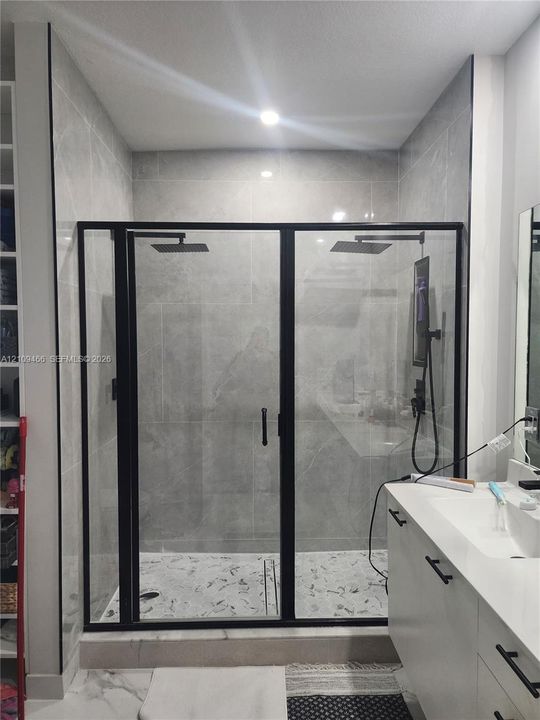 Walk-in shower featuring large-format tile surround, matte black metal-framed glass enclosure, dual overhead rainfall showerheads, and a patterned tile floor