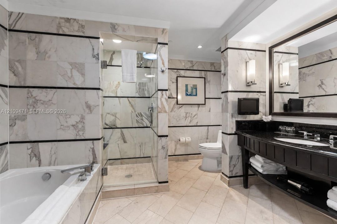 Bathroom featuring full-height natural stone wall tiling with horizontal accent banding, a glass-enclosed shower, a deep soaking tub, and a vanity with a dark stone countertop and integrated sink