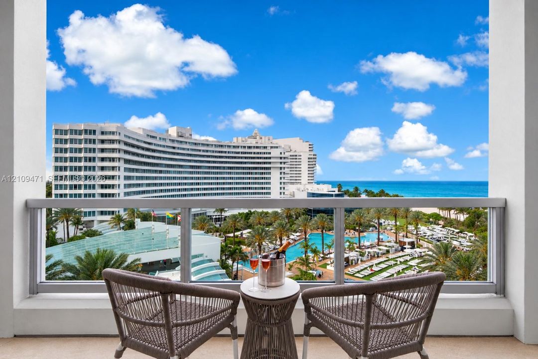 Covered balcony featuring a metal and glass railing, providing an elevated outlook over the ocean, sandy coastline, resort-style pools, and palm-lined grounds
