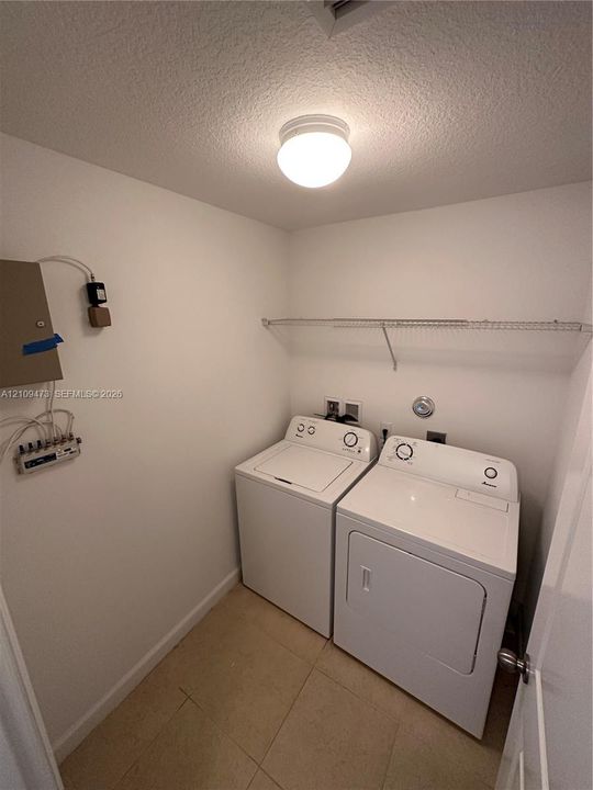 Utility space featuring large-format neutral tile flooring, a wall-mounted wire shelving unit, and a ceiling-mounted globe fixture