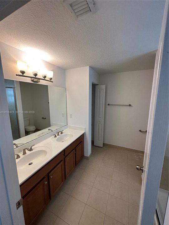 Bathroom featuring a dual-sink vanity with a solid-surface countertop, full-width wall mirror, wood-finish cabinetry, and ceramic tile flooring