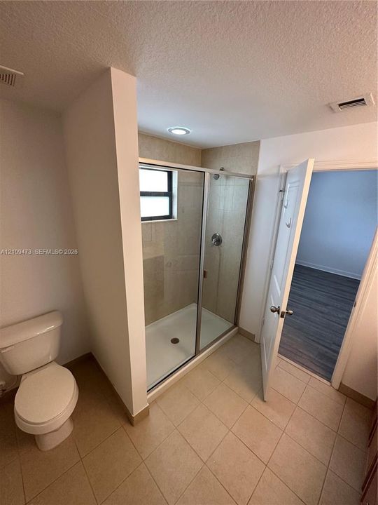Shower enclosure featuring glass doors, light-toned stone-finish tile surround, and recessed ceiling lighting, accompanied by a floor-mounted toilet and ceramic tile flooring