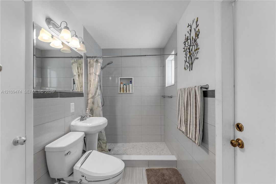 Bathroom featuring full-height gray wall tile, a walk-in shower with a recessed tiled niche, a pedestal sink, and a multi-bulb vanity light fixture