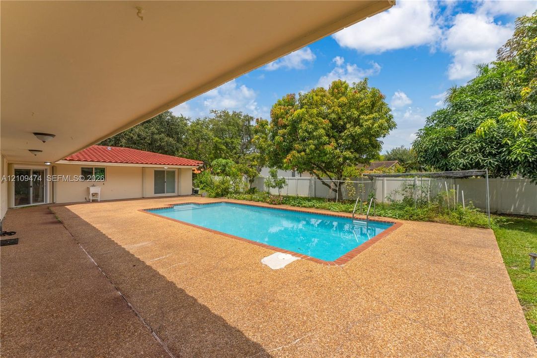 Rectangular in-ground swimming pool with a brick perimeter, extensive aggregate concrete patio, covered outdoor area, and white vinyl perimeter fencing