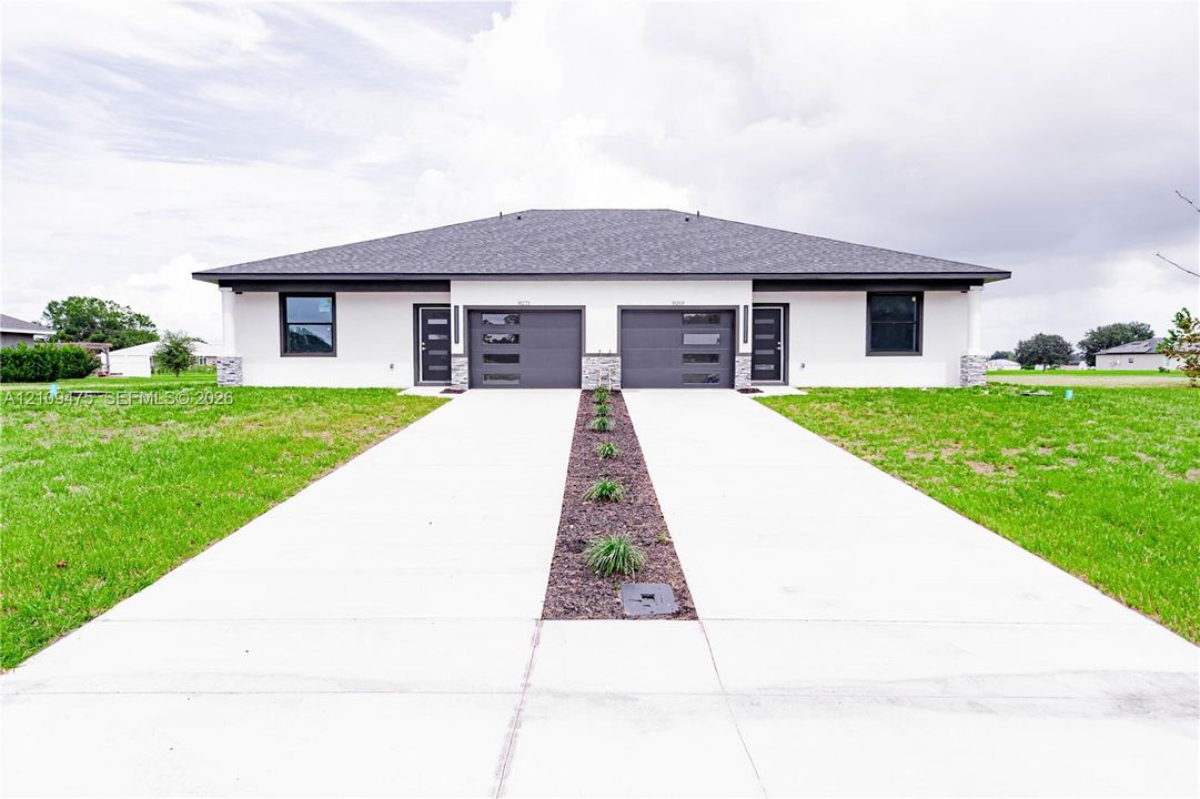 Modern exterior featuring white stucco finish, stone veneer accents, dual sectional garage doors with horizontal glass panels, and a split concrete driveway with a central rock garden border