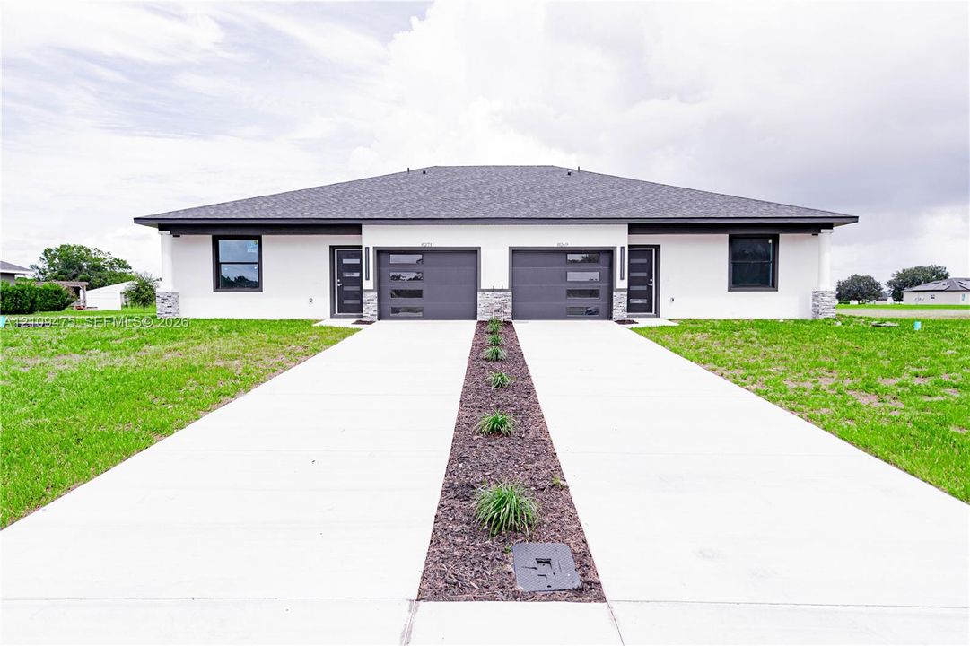 Contemporary duplex featuring a light-colored stucco exterior, dual attached garages with modern glass-paneled doors, stacked stone columns, and a concrete driveway with a central mulched garden bed