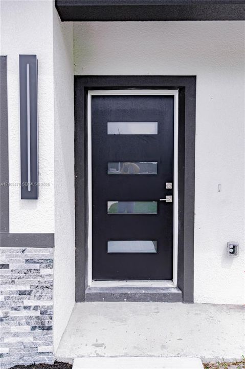 Modern black entry door featuring four horizontal glass inserts, brushed metal hardware, linear wall-mounted exterior lighting, and textured stone masonry detailing