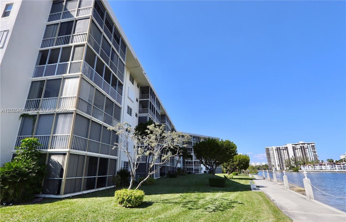 Multi-story building exterior featuring screened balconies, manicured lawn, concrete walkway, and waterfront bulkhead