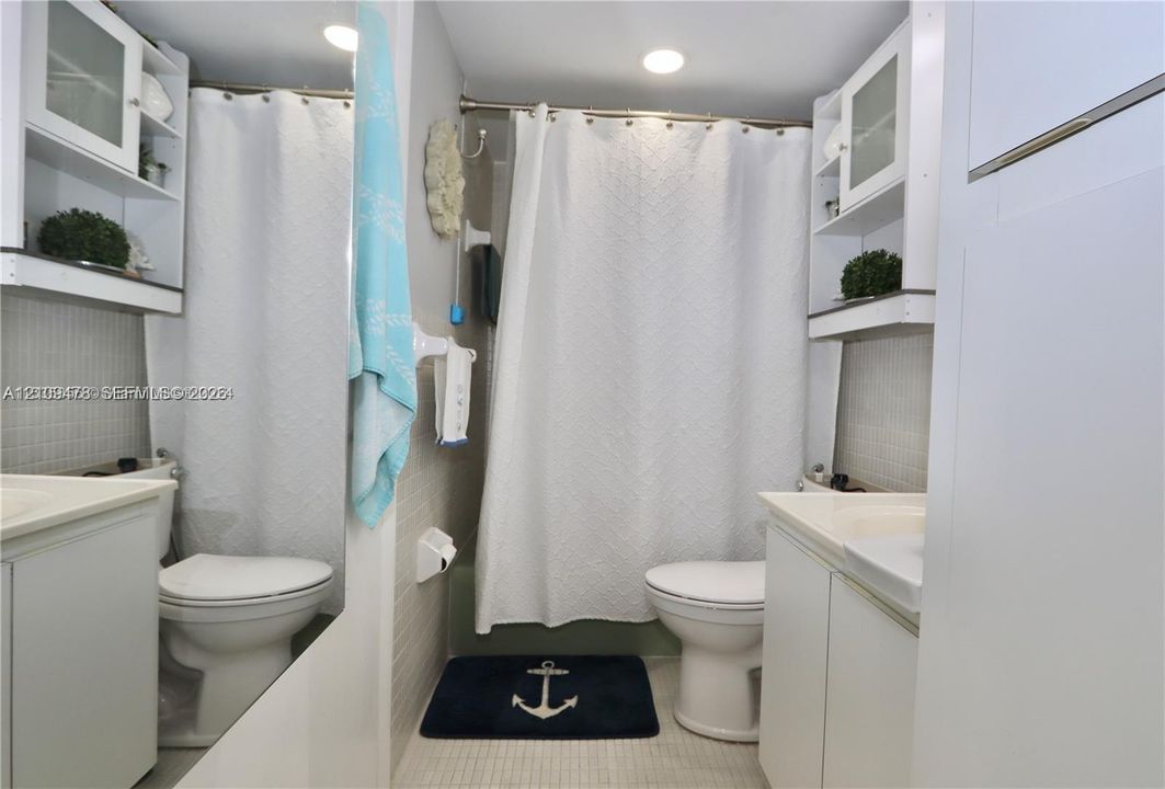 Full bathroom featuring dual white vanity cabinets, integrated white sinks, wall-mounted upper storage shelving, a tiled shower surround, and a large wall-mounted mirror