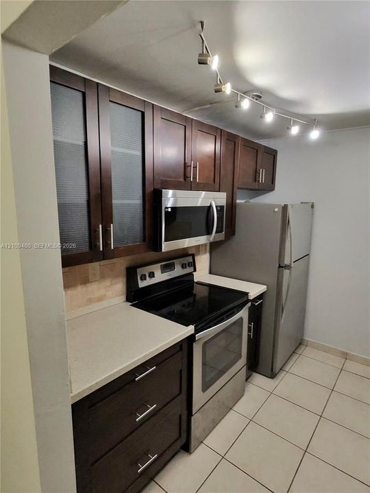 Kitchen featuring dark-finish cabinetry with glass-panel upper doors, stainless steel appliances, solid-surface countertops, tile flooring, and adjustable track lighting