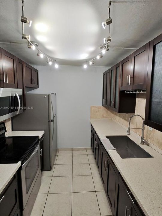 Kitchen featuring dark wood-finish cabinetry, solid surface countertops, stainless steel appliances, ceramic tile flooring, and adjustable track lighting