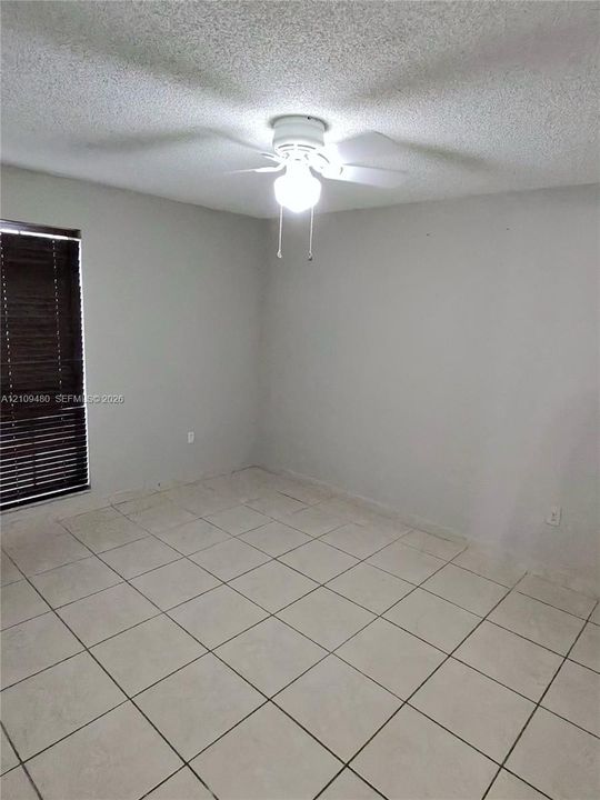 Neutral-toned interior room featuring light-colored tile flooring, a ceiling-mounted light fixture with a fan, and a window with horizontal slatted blinds