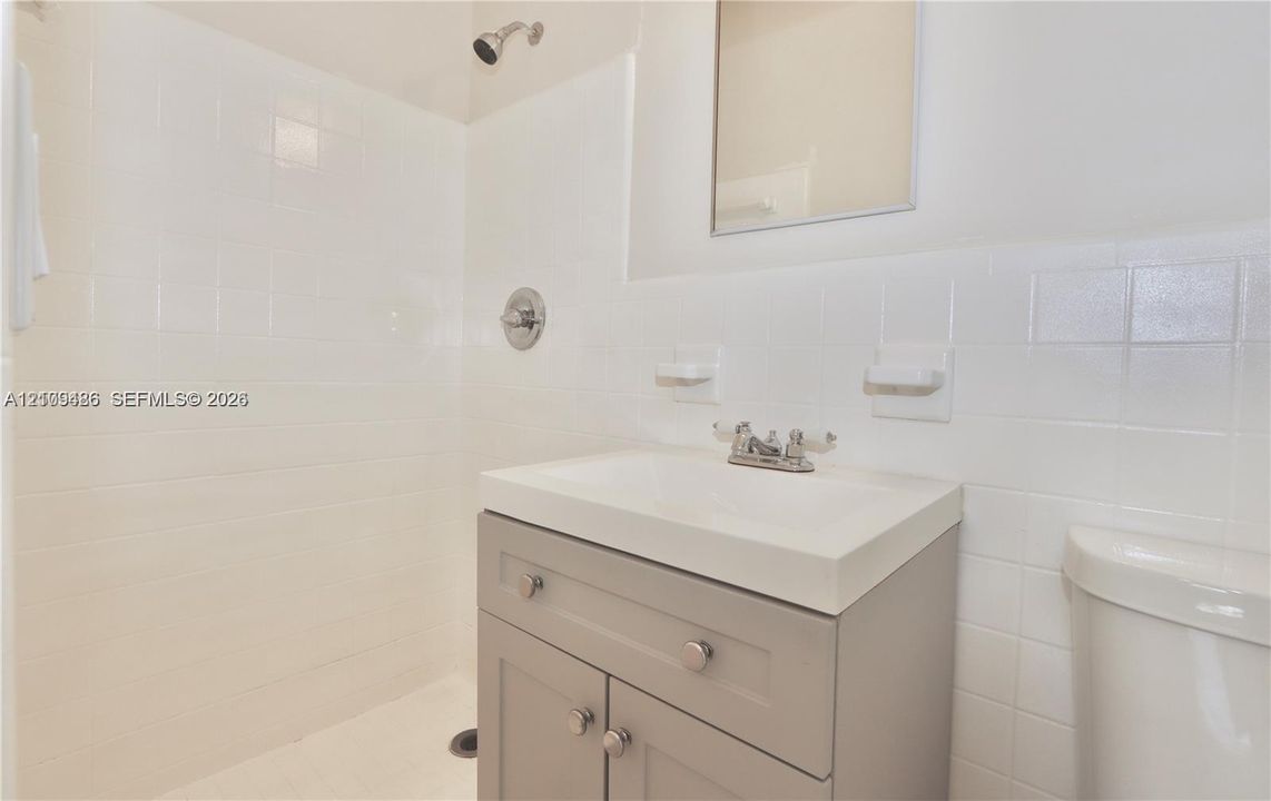 Bathroom featuring a grey vanity with a rectangular sink, wall-mounted mirror, and white tile surround in the shower area
