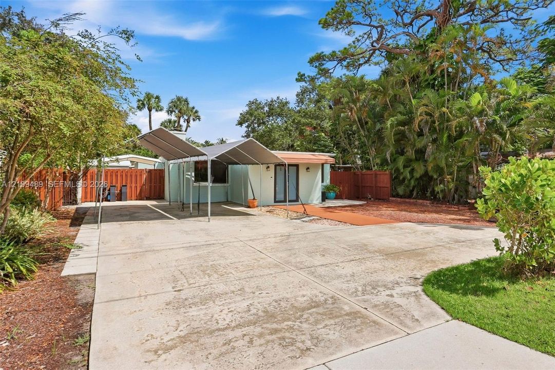 Exterior space featuring a concrete driveway, metal-framed carport, fenced perimeter, and mature landscape