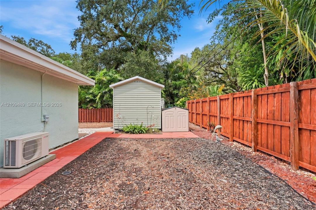 Rear exterior featuring a wood-style privacy fence, red paver pathways, dedicated storage structures, and mature tree canopy