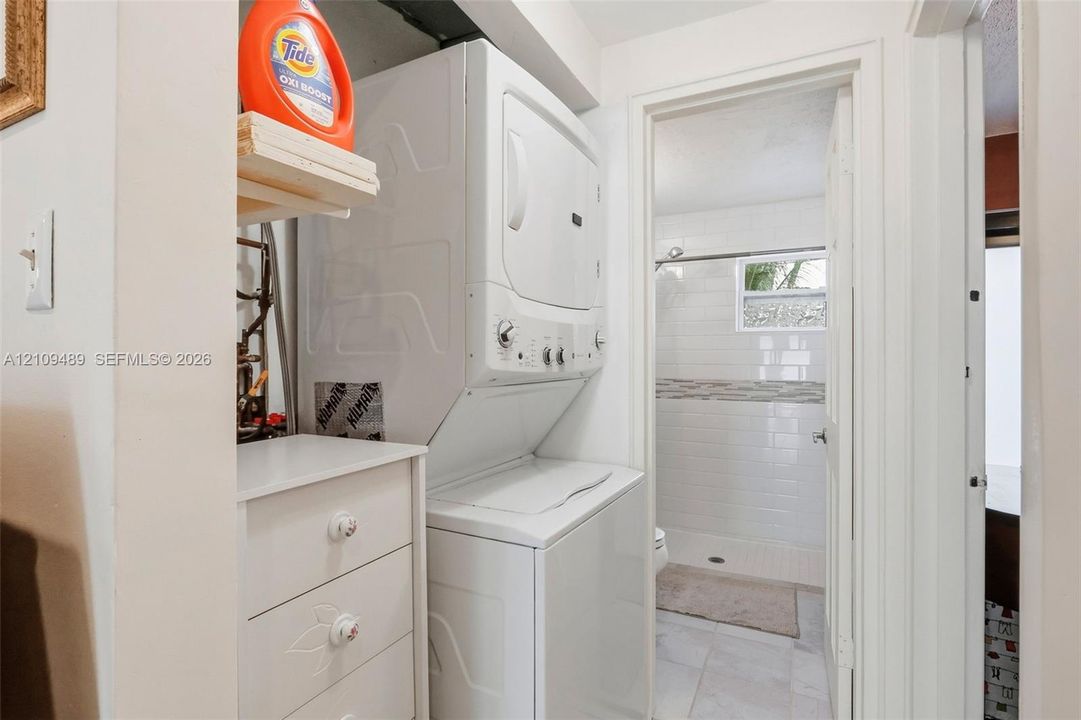 Stacked laundry unit with built-in storage cabinetry, white tile bathroom flooring, subway tile shower surround, and a framed window