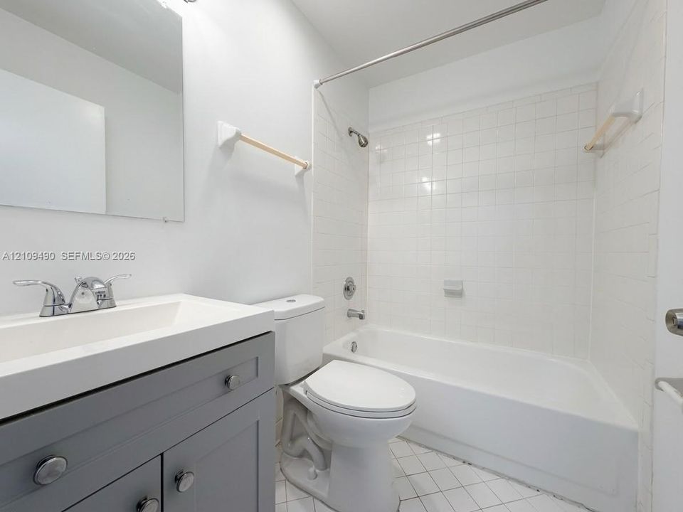 Bathroom featuring a grey vanity with a white countertop, wall-mounted mirror, white ceramic tile shower surround, and white tile flooring