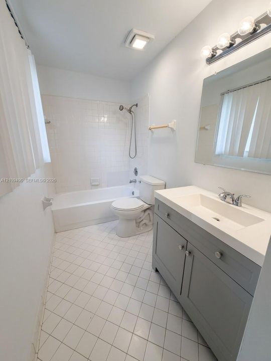 Gray vanity with integrated rectangular sink, solid surface countertop, tile-surround bathtub with shower head, and ceramic tile flooring