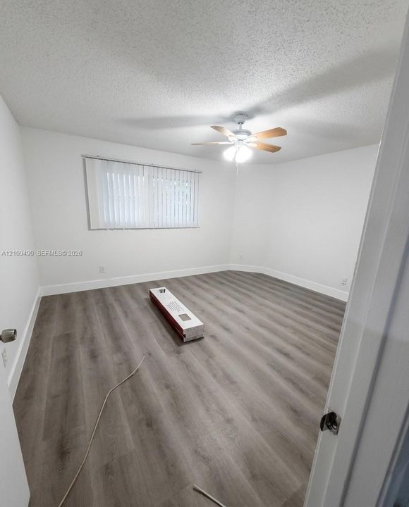 Neutral-toned interior space featuring wood-finish flooring, a centrally mounted ceiling fan with integrated lighting, and horizontal window blinds
