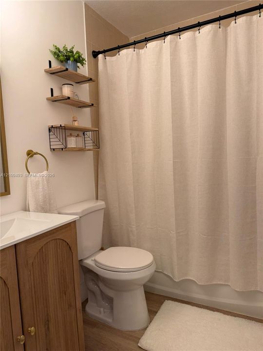 Bathroom featuring a wood-finish vanity with a white countertop, wall-mounted wood shelving, brushed-gold towel ring, and a shower with a black curtain rod
