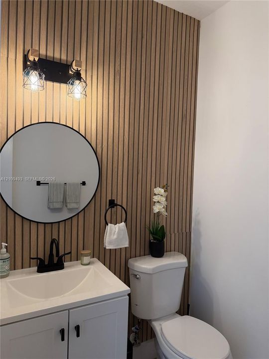 Vertical slat wood-finish accent wall featuring a round black-framed mirror, white vanity cabinet with matte black hardware, and two-light industrial-style wall sconce