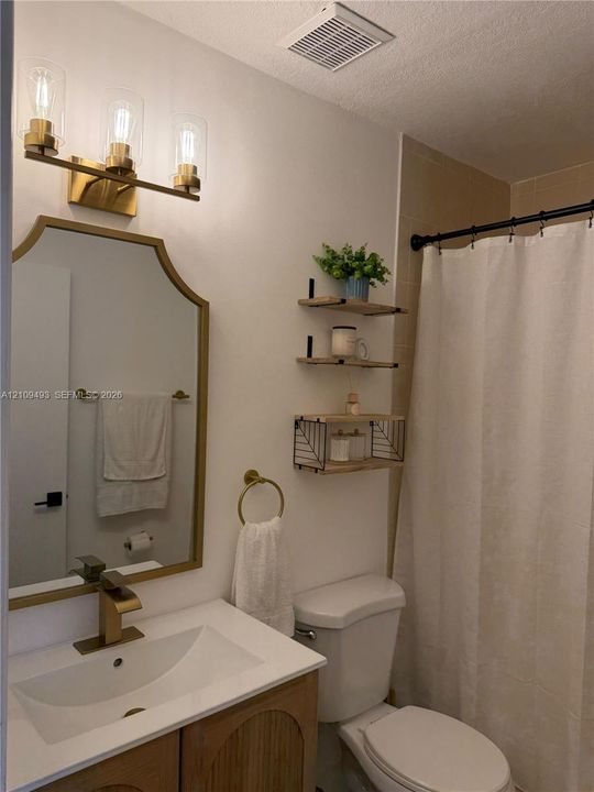 Vanity featuring a white countertop and gold-tone faucet, arched wall mirror with a gold-tone frame, three-light vanity fixture with clear glass shades, and wall-mounted shelving