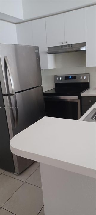 Kitchen featuring white cabinetry, stainless steel refrigerator, electric range with matching hood, solid-surface countertops, and tile flooring