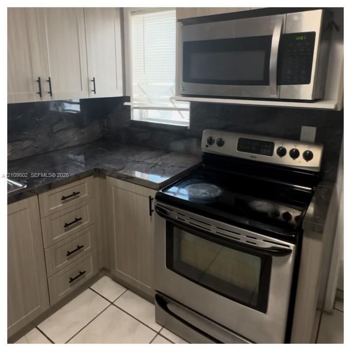 Light-toned cabinetry with matte black hardware, dark stone-finish countertops and backsplash, stainless steel electric range, and integrated microwave