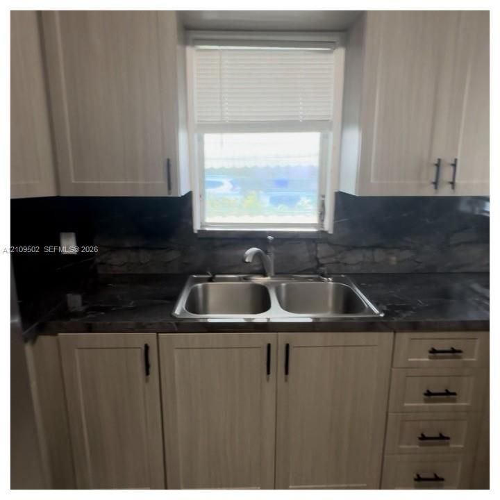 Light-toned cabinetry featuring matte black hardware, dark stone-patterned countertop, full-height matching backsplash, stainless steel dual-basin sink, and a gooseneck faucet
