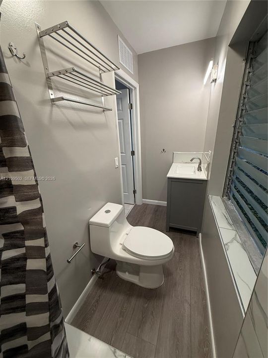 Bathroom featuring wood-finish flooring, a charcoal gray vanity with a square sink, a one-piece toilet, and a wall-mounted metal tiered shelving unit