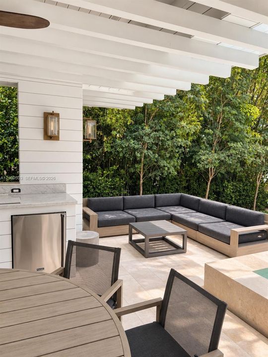 Covered patio featuring a white horizontal plank facade, built-in outdoor kitchen with stainless steel refrigeration, natural stone flooring, and wall-mounted brass-finish light fixtures