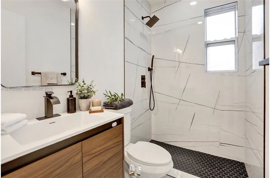 Wood-finish vanity with a rectangular sink, large-format tile shower surround featuring stone-style veining, matte black hardware, and black hexagonal floor tile