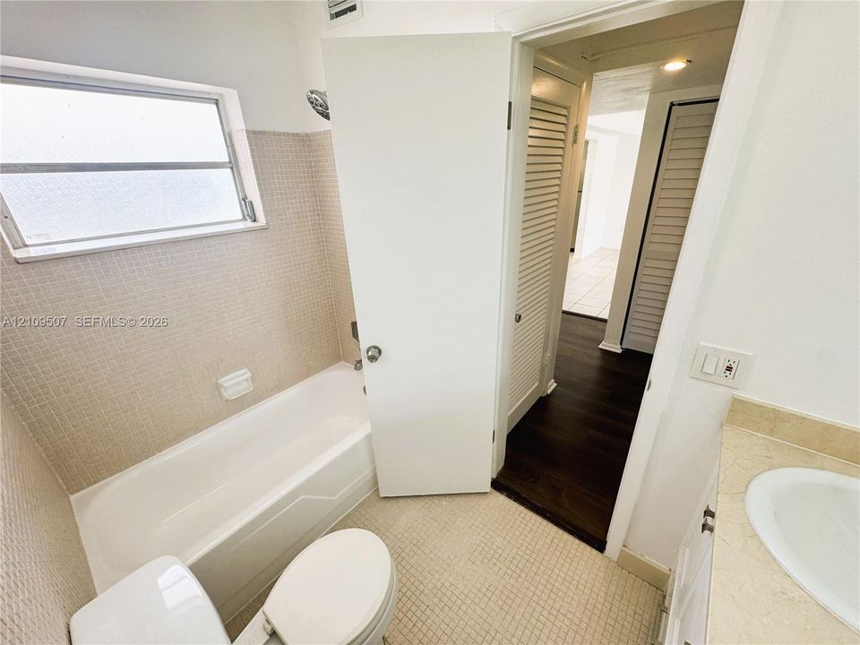 Bathroom featuring square mosaic wall tiling, a built-in bathtub with shower head, a white vanity with integrated oval sink, and louvered closet doors
