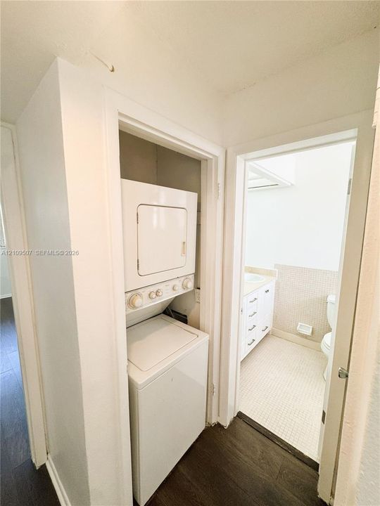 Dedicated laundry alcove with stacked white washer and dryer unit, wood-finish flooring, and doorway access to tiled bathroom featuring a white vanity cabinet and undermount sink
