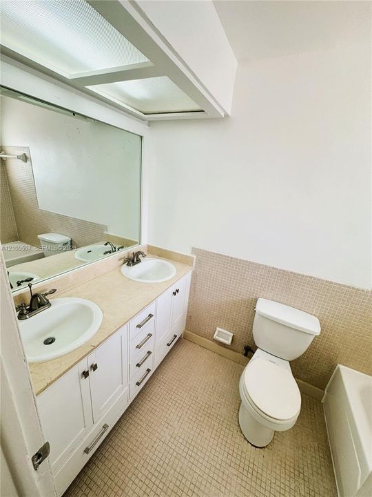 Dual-sink vanity with a stone countertop, inset sinks, white cabinetry, a large wall-mounted mirror, and tile wainscoting