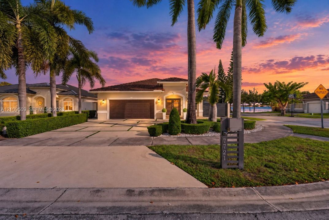 Stucco exterior with a tiled roof, multi-car garage, expansive concrete driveway with geometric lawn inlays, and mature palm tree landscaping