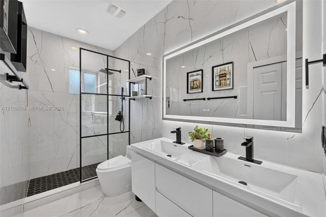 Modern bathroom featuring large-format veined stone tile walls, a dual-sink vanity with matte black faucets, a frameless glass-enclosed shower with black metal grid framing, and a large backlit vanity mirror