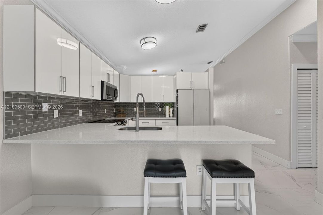 Kitchen featuring high-gloss white cabinetry, white stone-finish countertops, stainless steel appliances, dark grey subway tile backsplash, and light-toned tile flooring